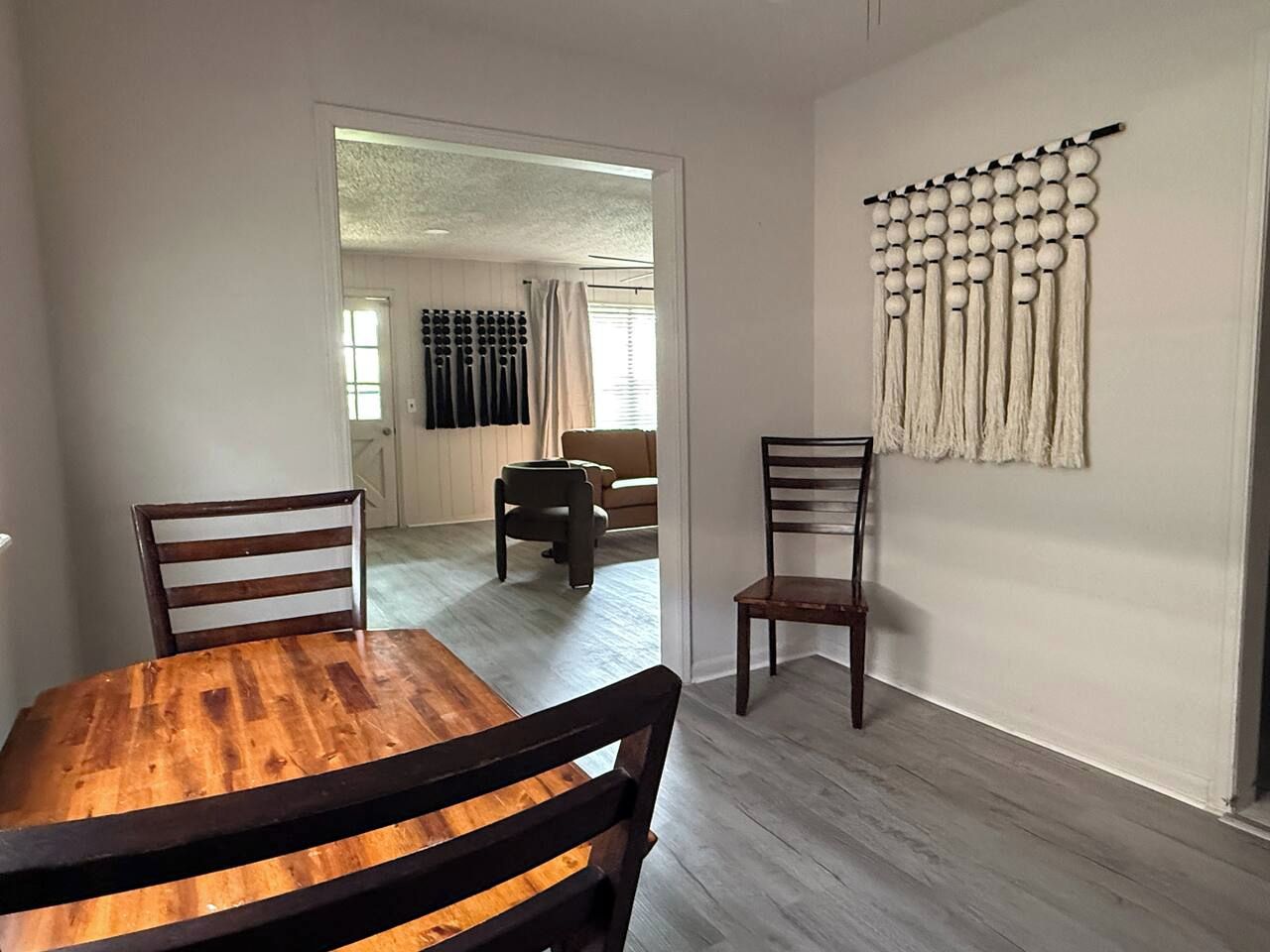 A dining area with a wooden table and chairs, a wall-mounted decorative piece, and a view into another room with a sofa and curtains.