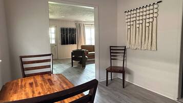 A dining area with a wooden table and chairs, a wall-mounted decorative piece, and a view into another room with a sofa and curtains.