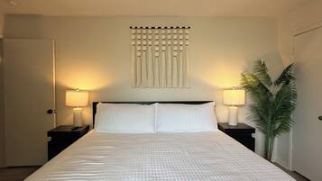 A bedroom with a large bed, two bedside tables with lamps, a wall-mounted light fixture, and a potted plant.