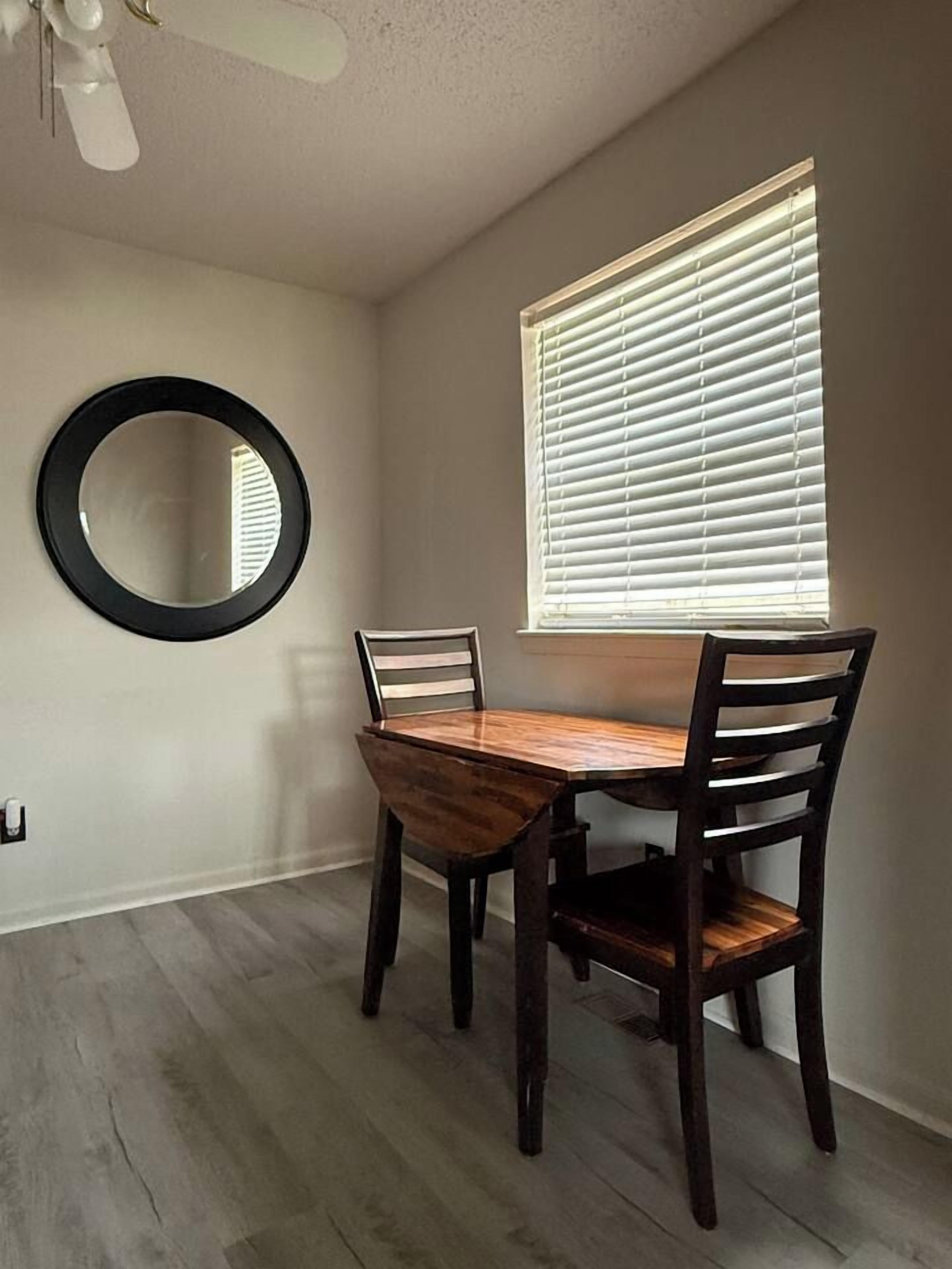 A dining area with a wooden table and chairs, a round mirror on the wall, and a window with blinds.