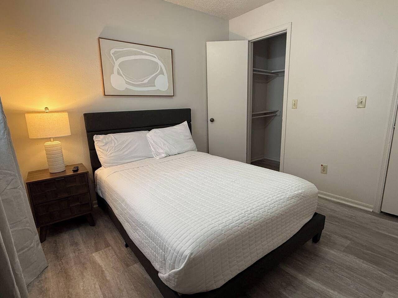 A bedroom with a bed, a nightstand, a lamp, a closet, and a wall-mounted artwork.