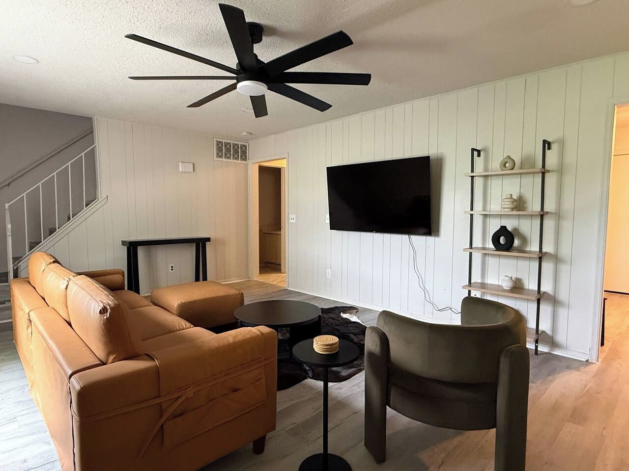 A living room with a sofa set, a coffee table, a chair, a flat-screen TV mounted on the wall, and a ceiling fan.