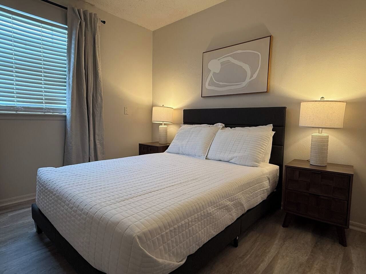 A neatly made bed with a quilted cover, two bedside tables with lamps, a wall-mounted abstract artwork, and a window with curtains.