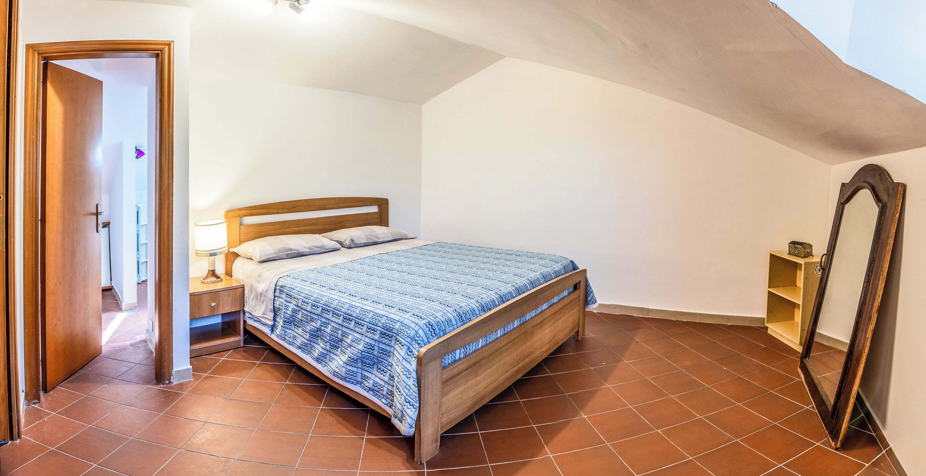 Un dormitorio con cama de madera, mesita de noche, lámpara de pie, espejo y suelo de baldosas.