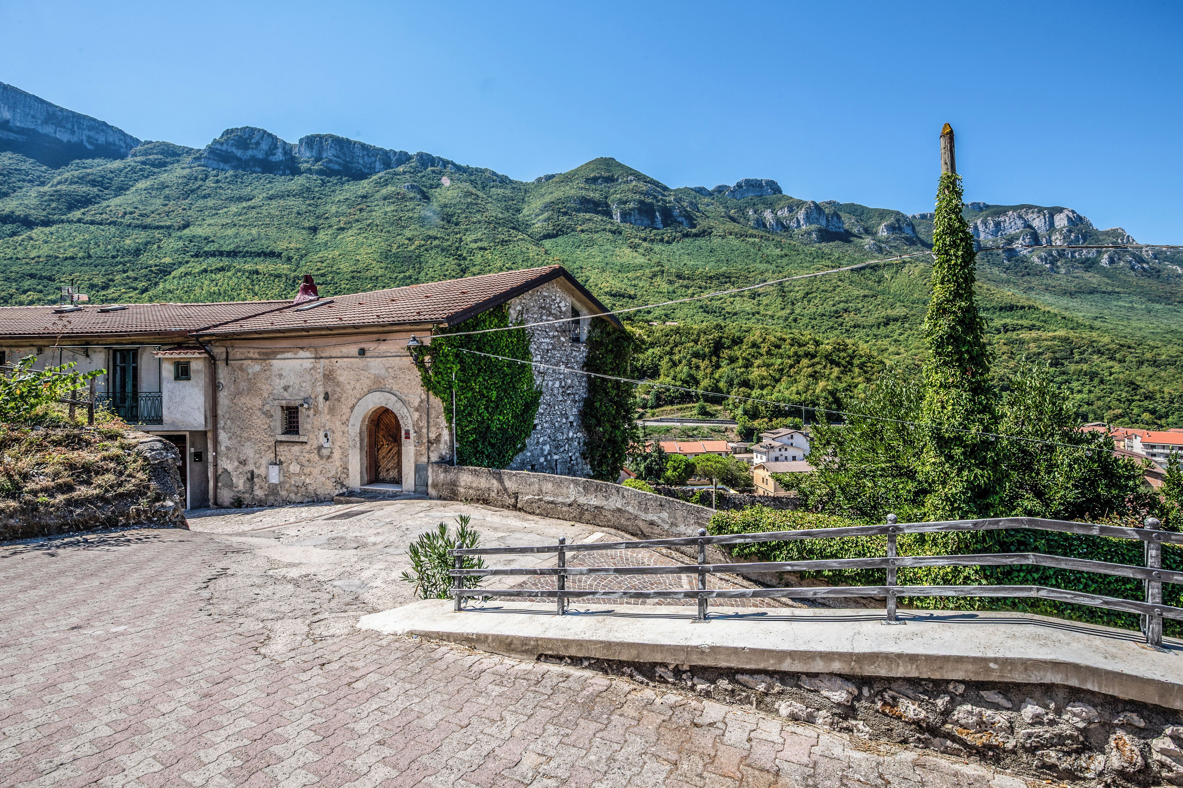 A rustic stone building with a tiled roof, a paved courtyard, and a view of a mountainous landscape.