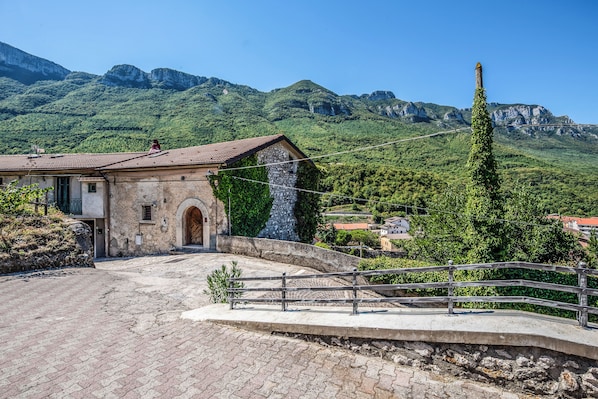 A rustic stone building with a tiled roof, a paved courtyard, and a view of a mountainous landscape.
