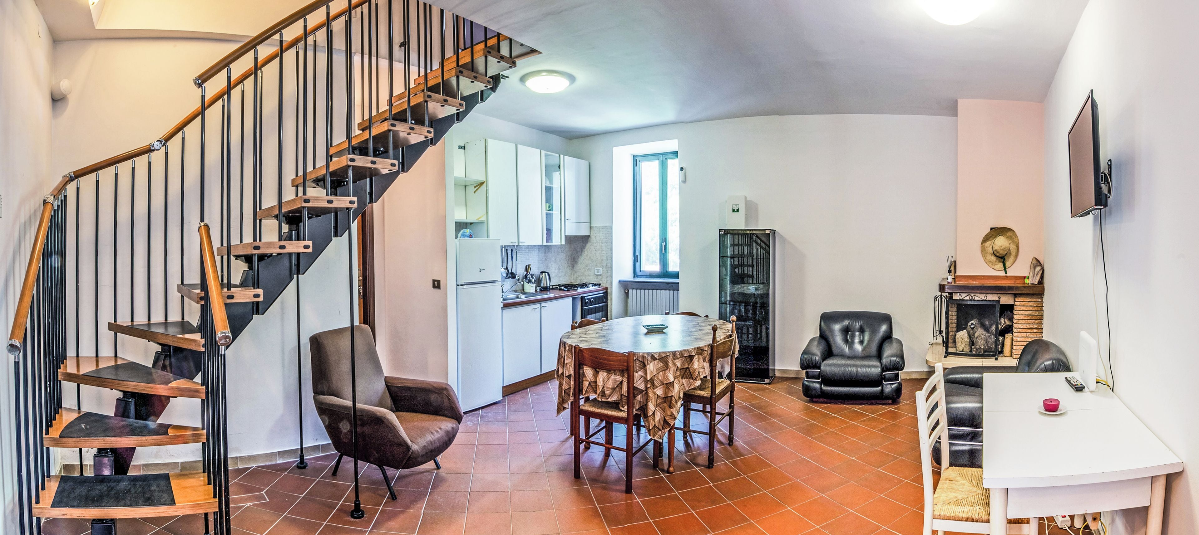 A spacious living area with a wooden staircase, a dining table with chairs, a kitchen in the background, and a fireplace.
