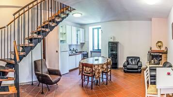 A spacious living area with a wooden staircase, a dining table with chairs, a kitchen in the background, and a fireplace.