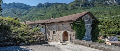 A stone building with a tiled roof, an arched entrance, and a view of green mountains in the background.