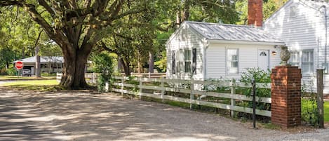 A white house with a brick chimney, surrounded by a white picket fence, and a large tree in the foreground.