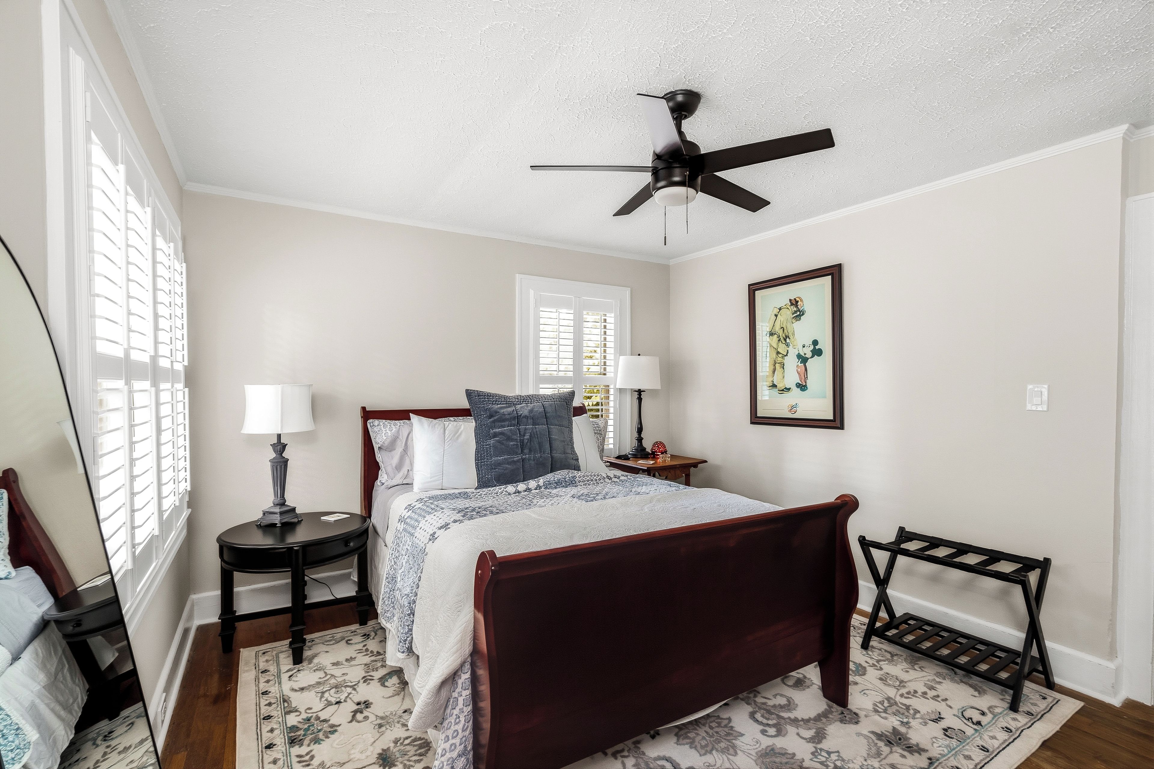 A bedroom with a bed, bedside tables, a ceiling fan, a window with blinds, and a framed picture on the wall.