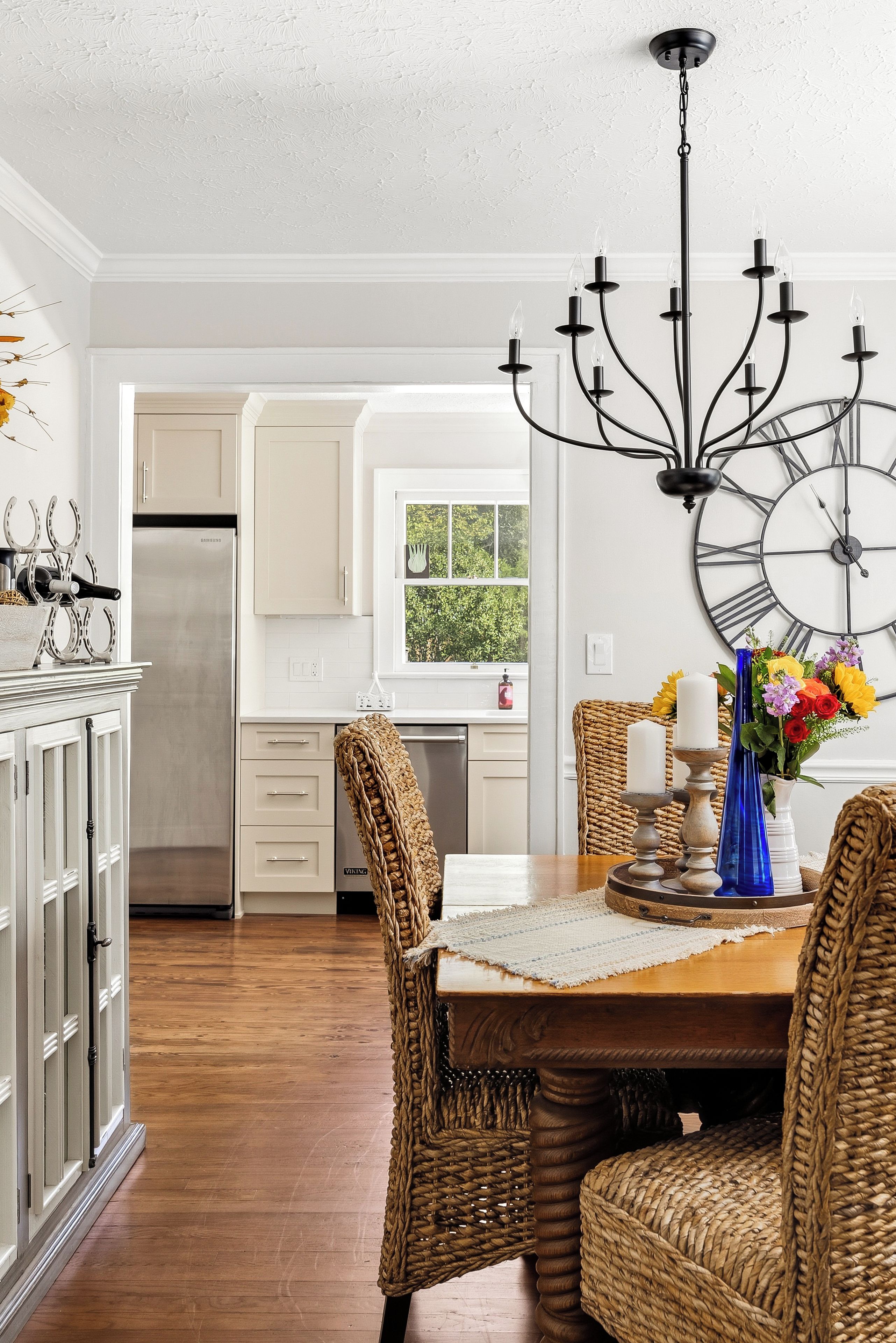 A kitchen with white cabinets, a stainless steel refrigerator, a dining table with a woven table runner, and a wall-mounted clock.