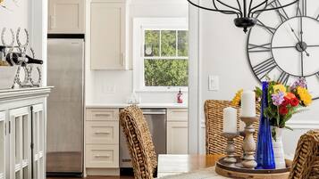 A kitchen with white cabinets, a stainless steel refrigerator, a dining table with a woven table runner, and a wall-mounted clock.