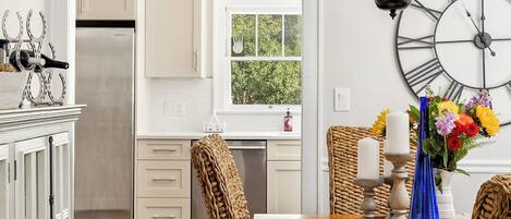 Una cocina con armarios blancos, un refrigerador de acero inoxidable, una mesa de comedor con camino de mesa tejido y un reloj montado en la pared.