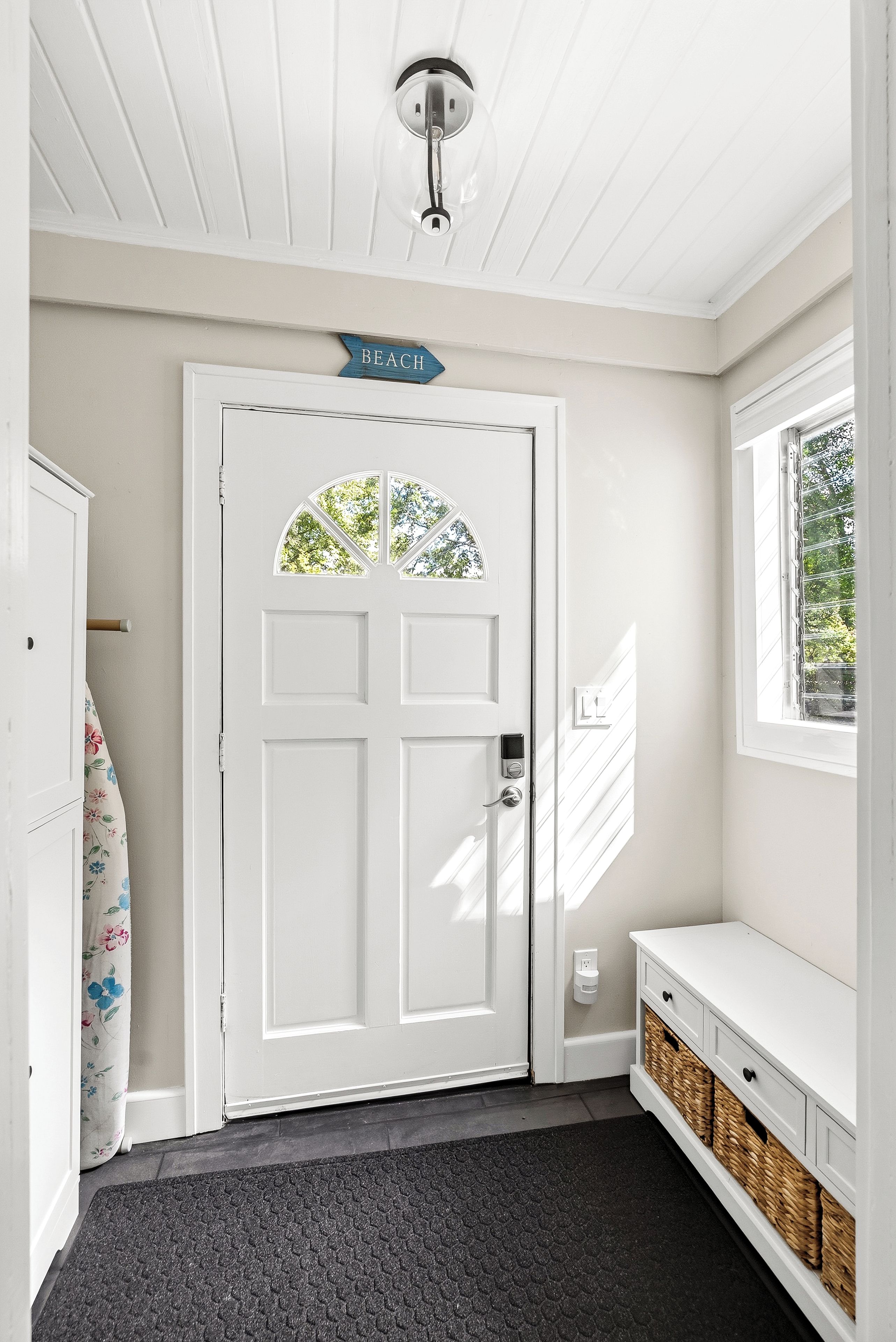 A white door with a 'BEACH' sign above it, a window with a view of trees, and a bench with baskets underneath.