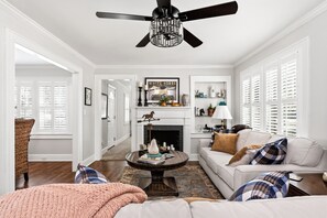 A well-lit living room with a fireplace, a ceiling fan, and a cozy seating area. - The Cottage on Boundary (Aiken)