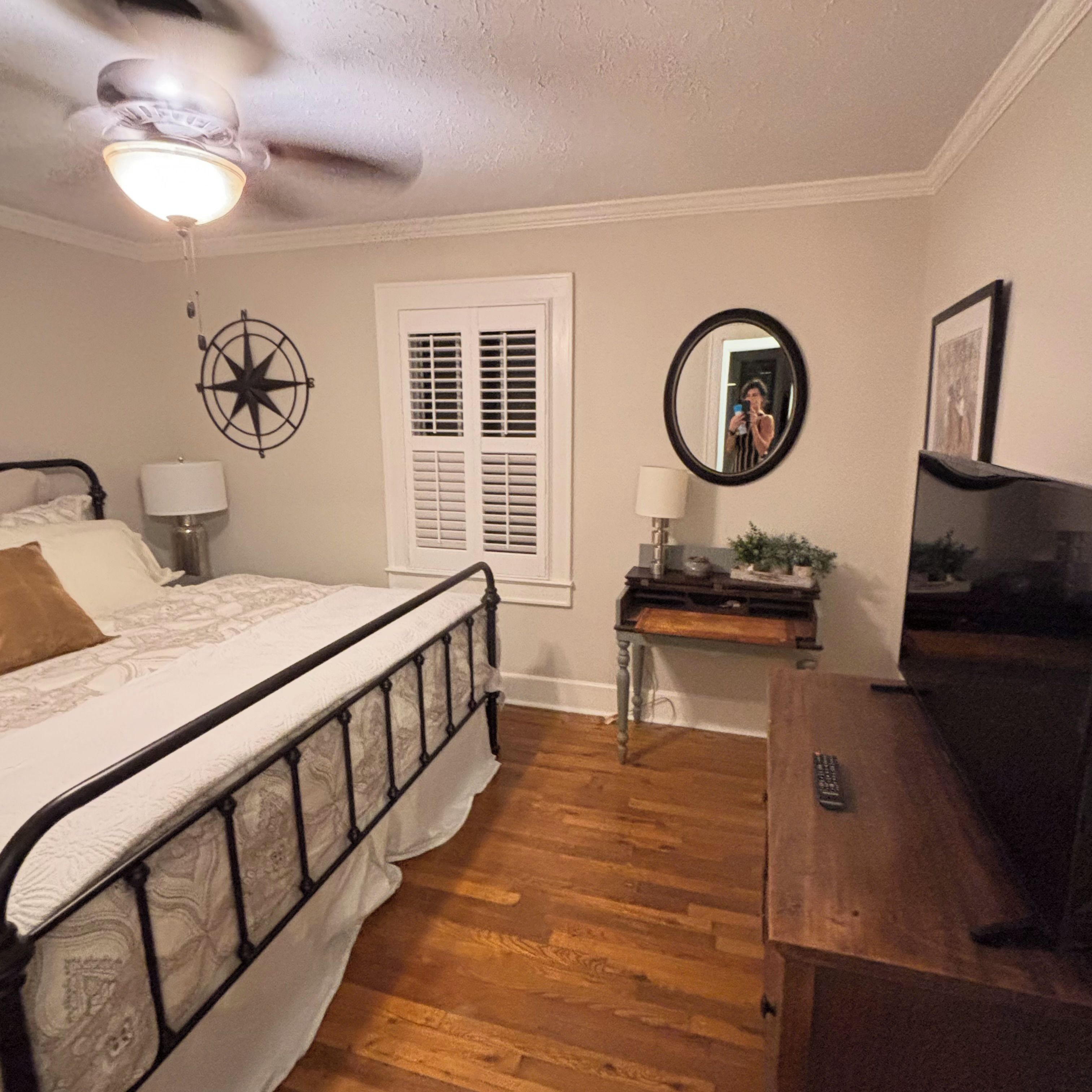 Un dormitorio con cama, escritorio, espejo, ventilador de techo y ventana con persianas.