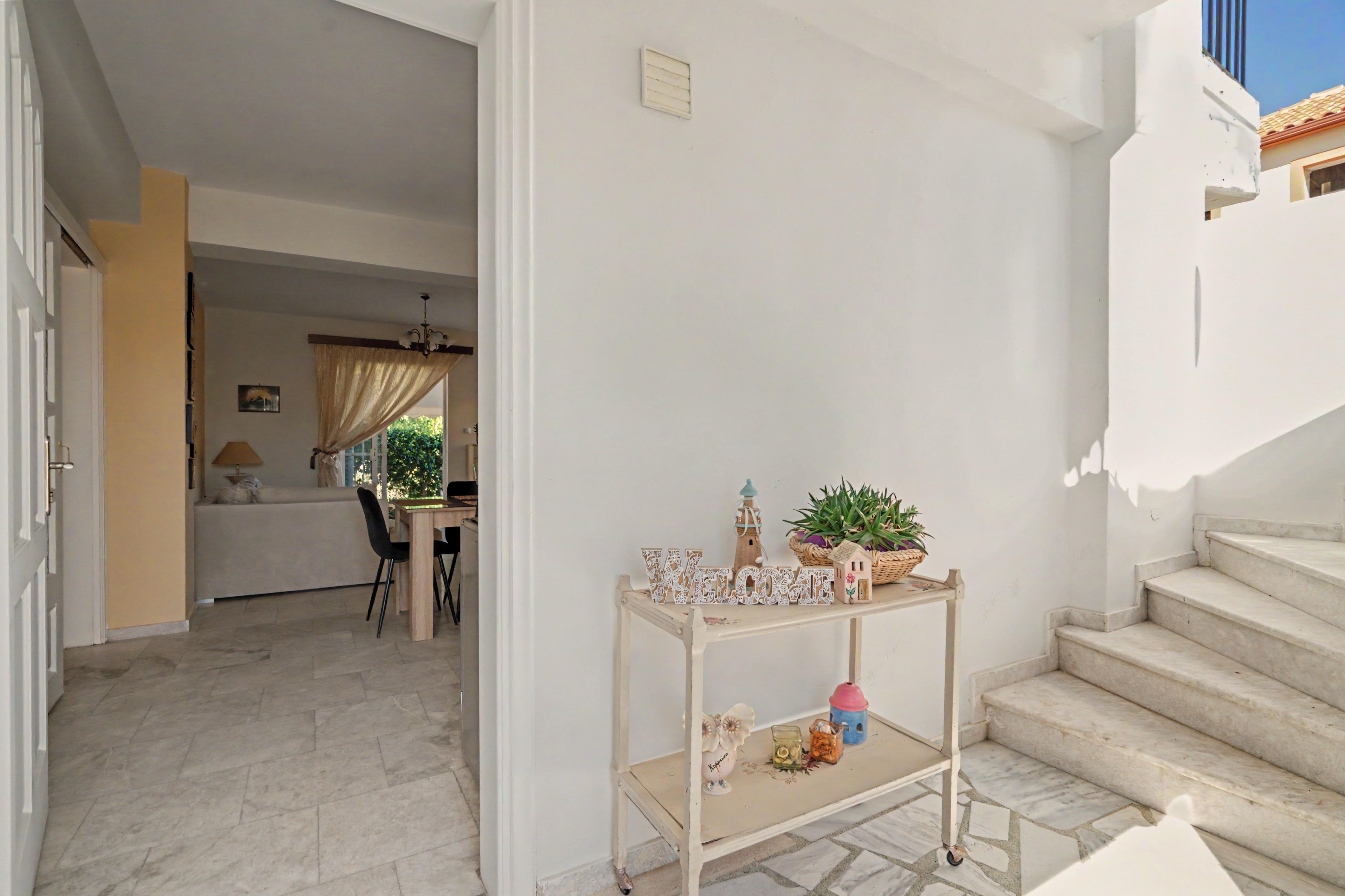 Un intérieur épuré, aux murs blancs, avec une table en bois où sont exposés des objets décoratifs, un escalier et une vue sur un coin repas.