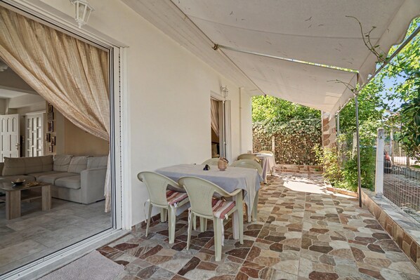 A covered outdoor seating area with a table and chairs, a sofa, and a view of the garden.