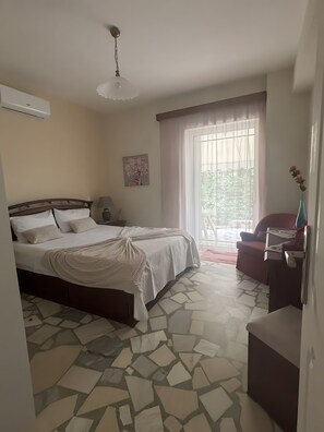 A bedroom with a bed, a nightstand, a lamp, a painting, a window with curtains, and a red chair. - Blossom Beachfront House with Private Terrace, Wi-Fi, and Air Conditioning (Κουρούτα)
