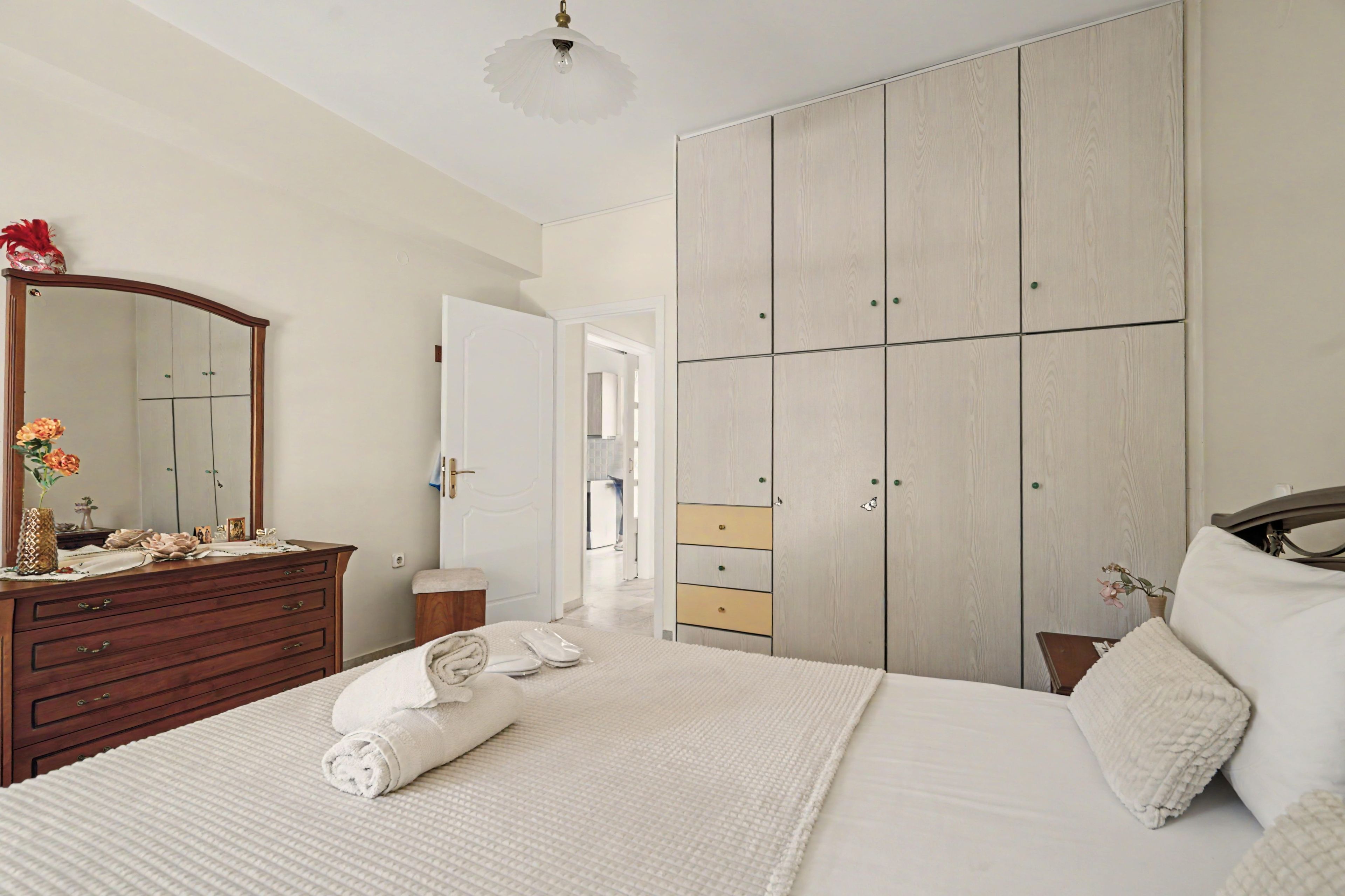 Une chambre avec une grande armoire, une commode en bois, un lit avec une literie blanche et un miroir agrémenté d’un vase de fleurs.