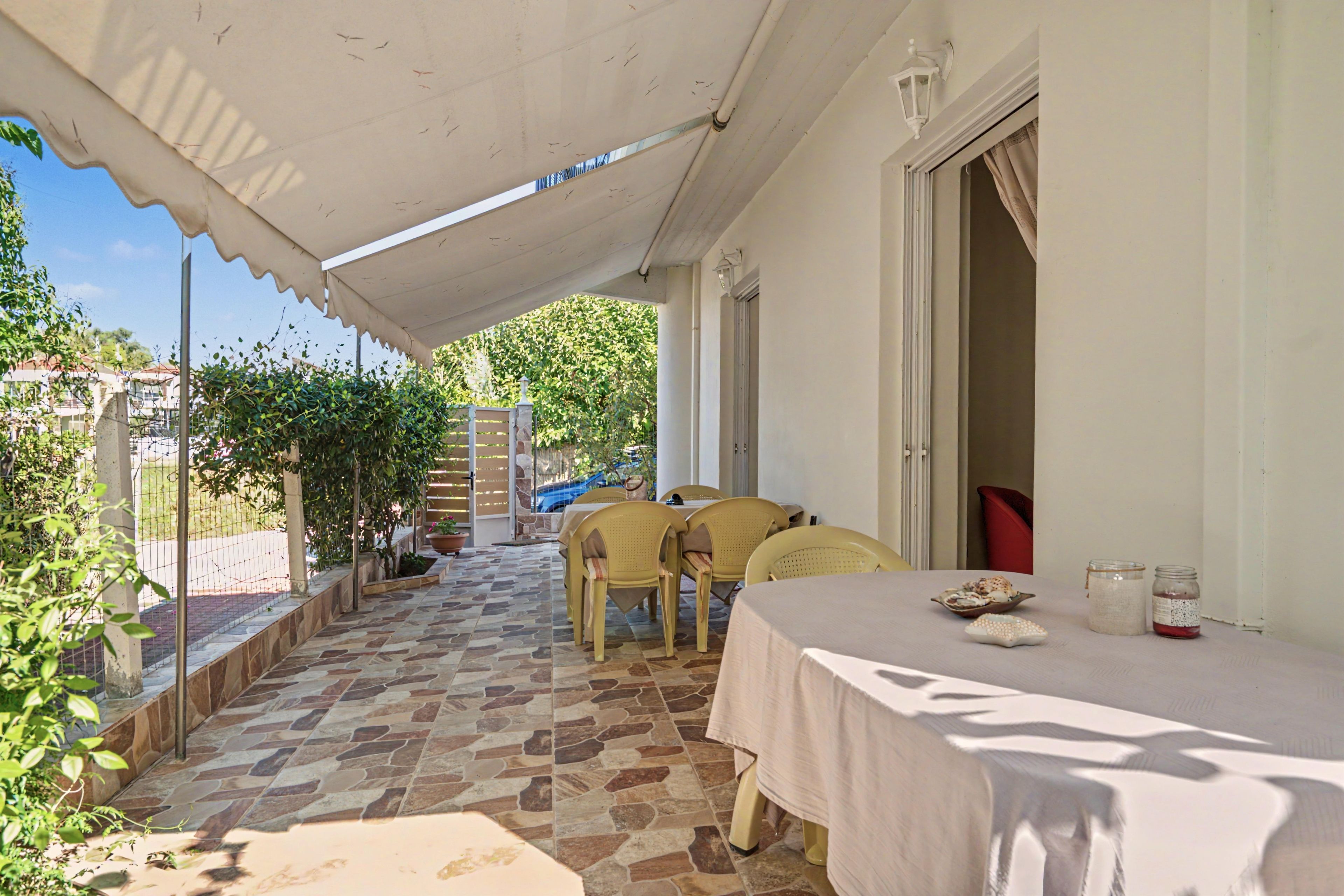 A covered outdoor dining area with stone flooring, a table with chairs, and a view of a garden.