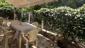 Eine Terrasse im Freien mit Tisch und Stühlen, einem überdachten Bereich und einem Garten mit blühenden Pflanzen.