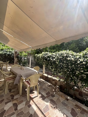An outdoor patio with a table and chairs, a covered area, and a garden with flowering plants. - Blossom Beachfront House with Private Terrace, Wi-Fi, and Air Conditioning (Κουρούτα)