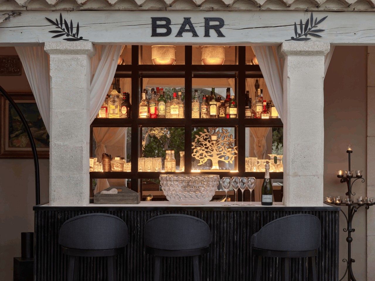 Een bar met een 'BAR' bord erboven, planken met flessen, een decoratieve boom en een kandelaar.