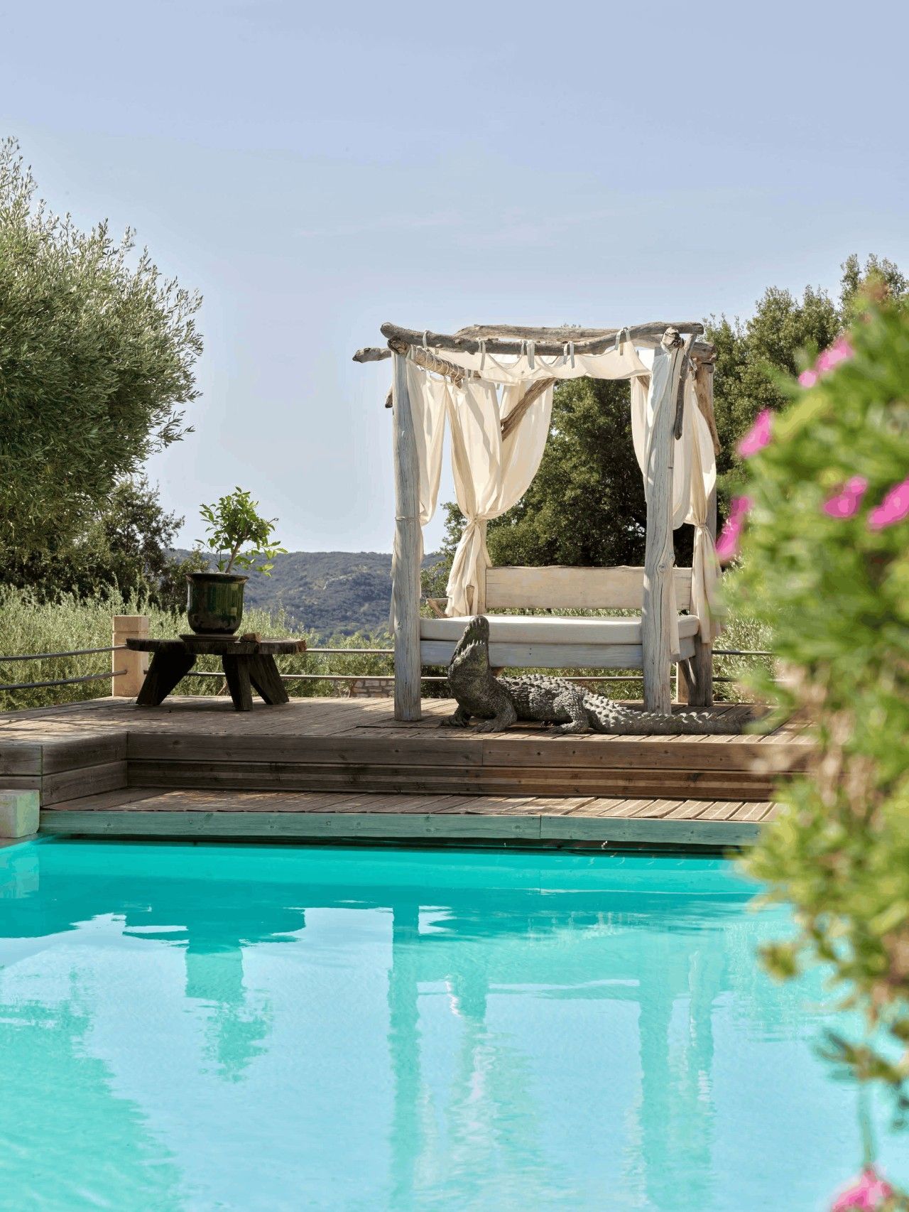 A pool with a wooden deck, a gazebo with white curtains, and a crocodile statue.
