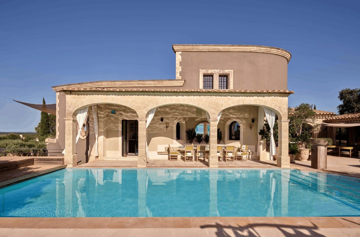 A poolside area with a pool, a covered seating area with chairs, and a building with arches.