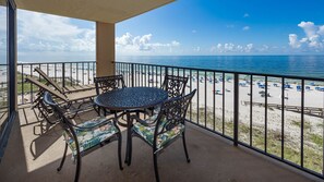 A balcony with a round table and chairs, overlooking a beach with umbrellas and people. - Phoenix I 1065 2 Bedroom Condo (Orange Beach)