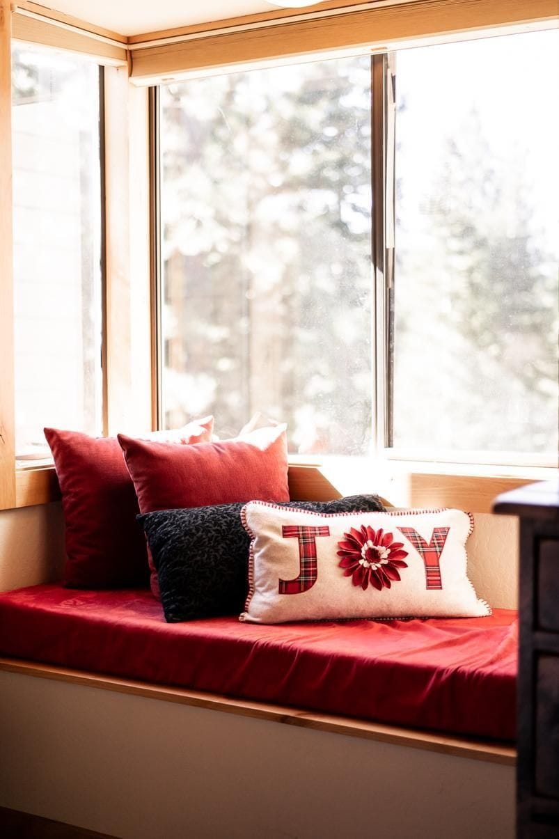 Asiento junto a la ventana con un cojín rojo, una almohada estampada y otra con la palabra 'JOY' bordada.
