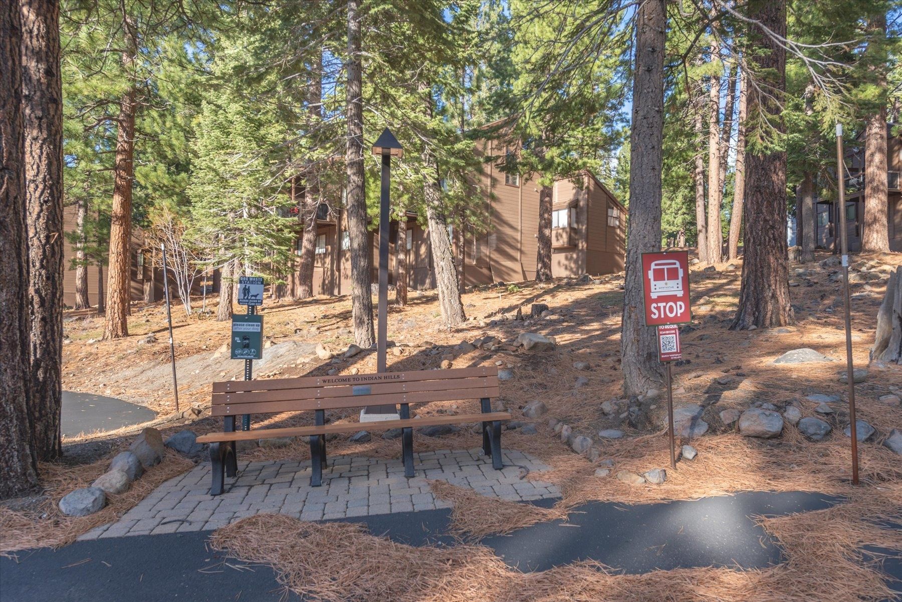 A bench in a forested area with a stop sign and a welcome sign to Indian Hills.