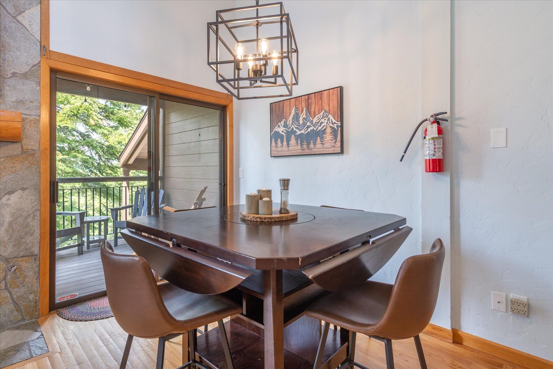 Un comedor con una mesa de madera, cuatro sillas, una lámpara colgante, un extintor y un cuadro en la pared.
