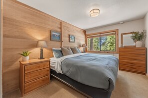 Ein Schlafzimmer mit Bett, Nachttischen, einer Kommode, einem Fenster mit Blick auf Bäume und Wandbildern.