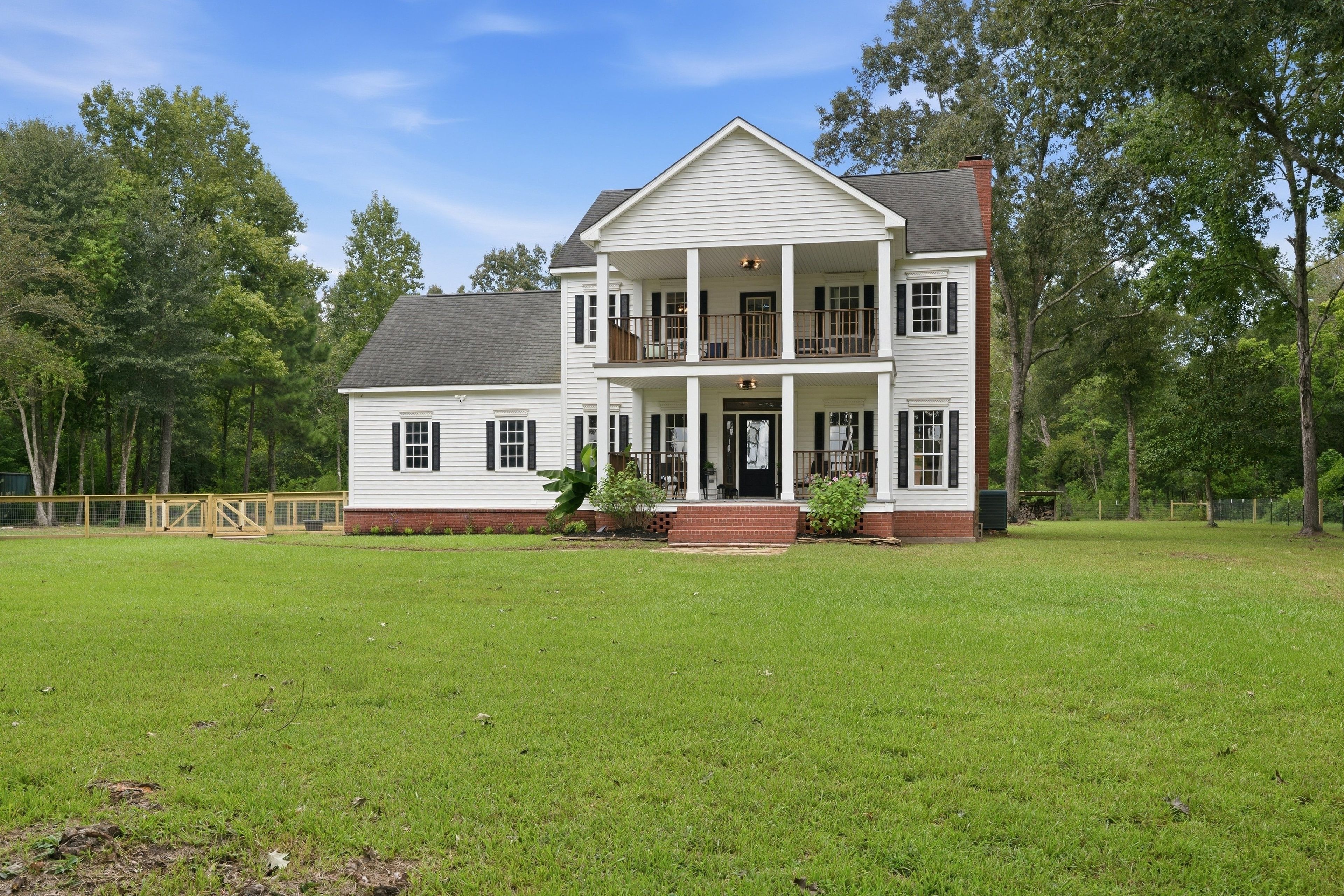 A two-story house with a front porch, a fenced yard, and trees in the background.