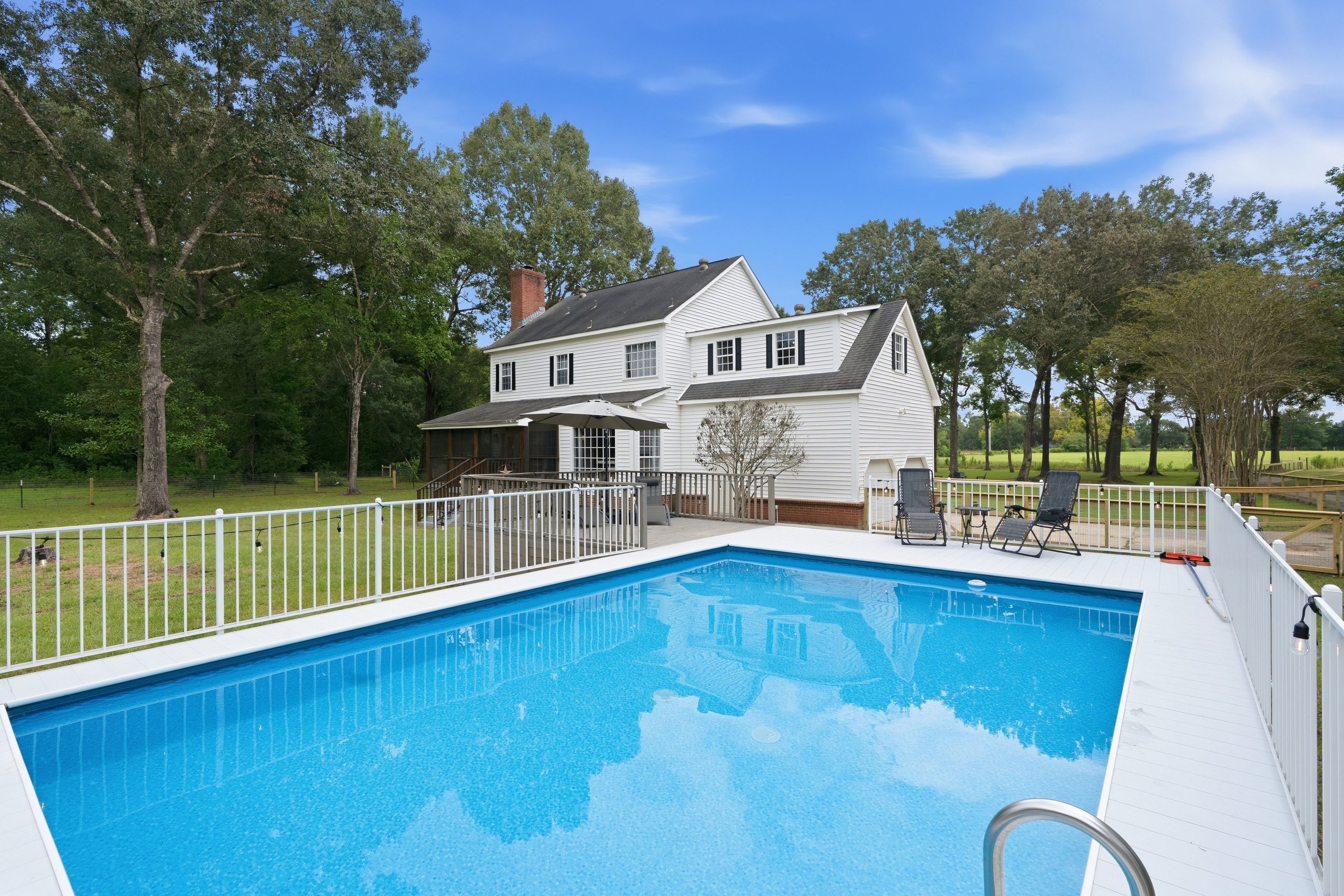 A swimming pool with a white deck, surrounded by a white fence, and a house with a chimney in the background.