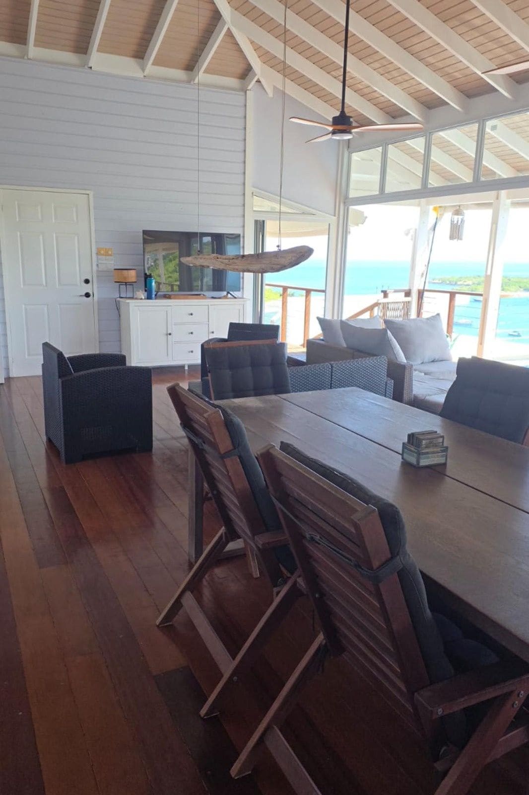 Uma sala de jantar com mesa e cadeiras de madeira, cozinha com armário branco e vista para o oceano.