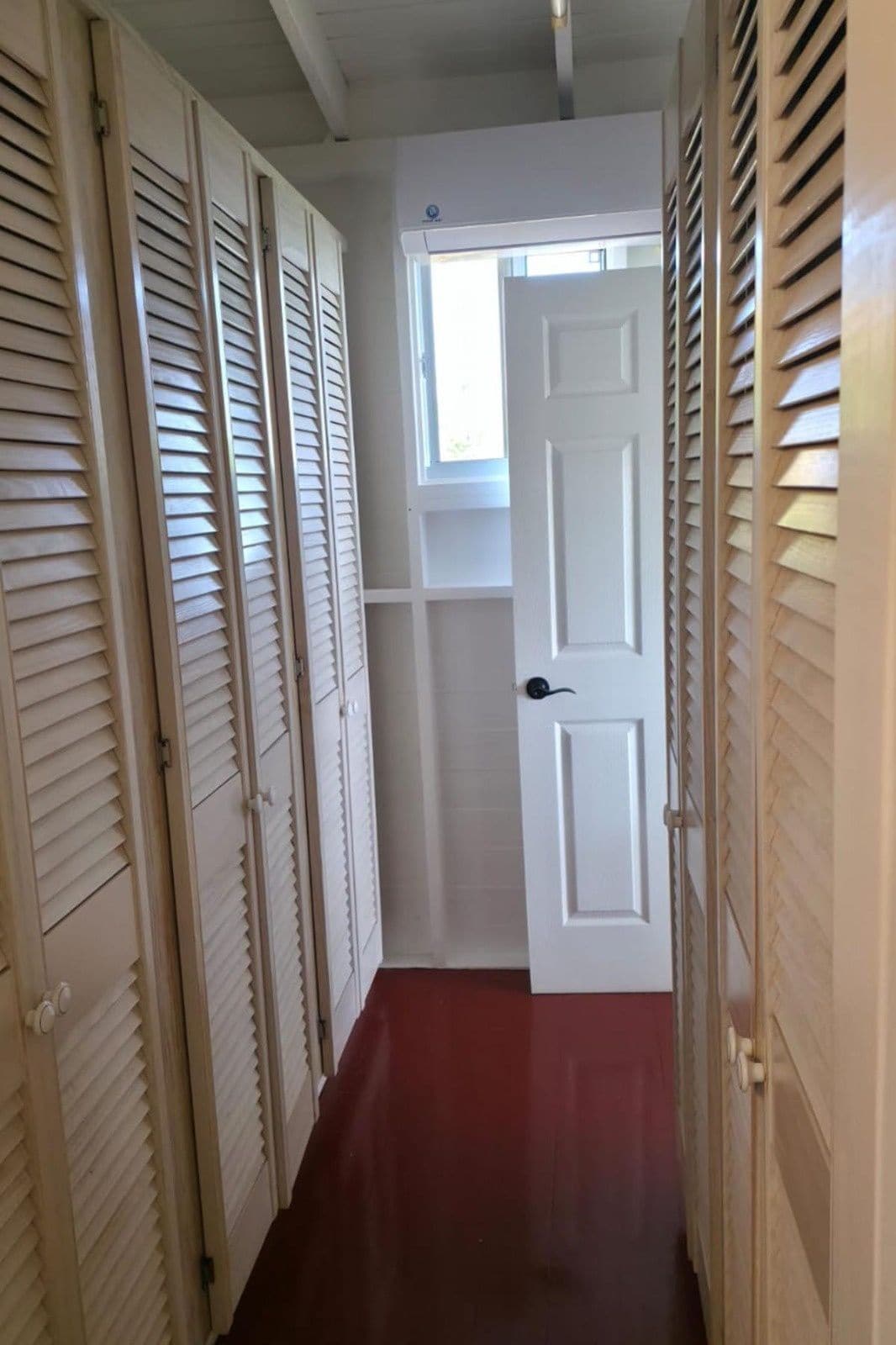 A narrow hallway with a red floor, white doors, and a window at the end.