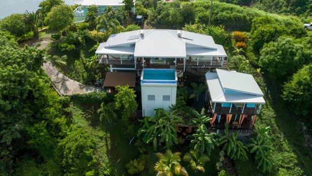 Uma casa grande, com vários andares, cercada por vegetação tropical densa, com uma estrutura azul no telhado em destaque.