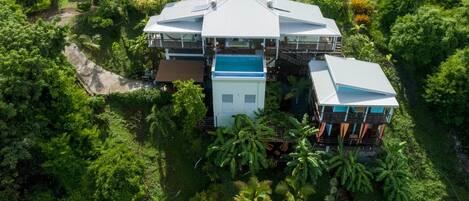 Une grande maison à étages, entourée d’une végétation tropicale luxuriante, avec une structure remarquable au toit bleu qui se dresse au centre.