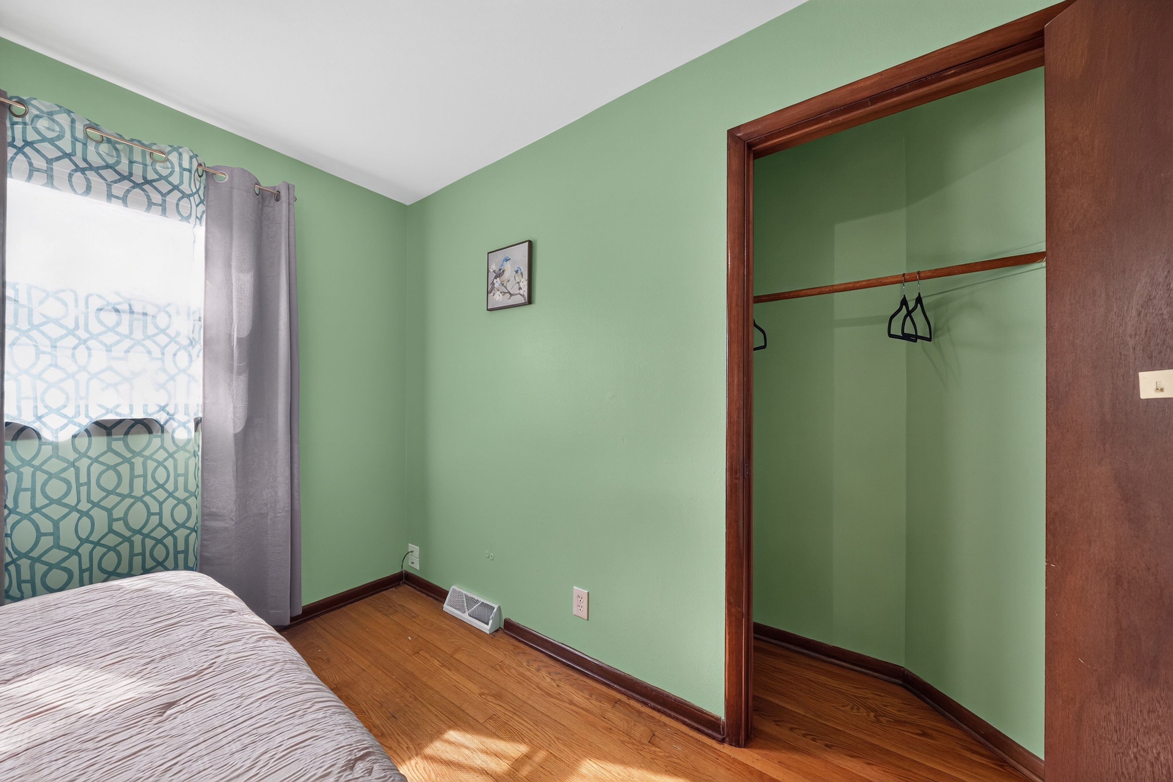 Une pièce avec un lit, une fenêtre ornée de rideaux à motifs, un tableau accroché au mur et une armoire en bois.
