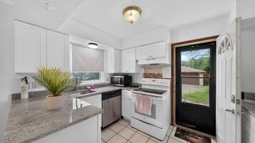 Cocina compacta con armarios blancos, microondas, lavavajillas y estufa. Hay una planta en maceta en la encimera y una ventana con vistas al exterior.