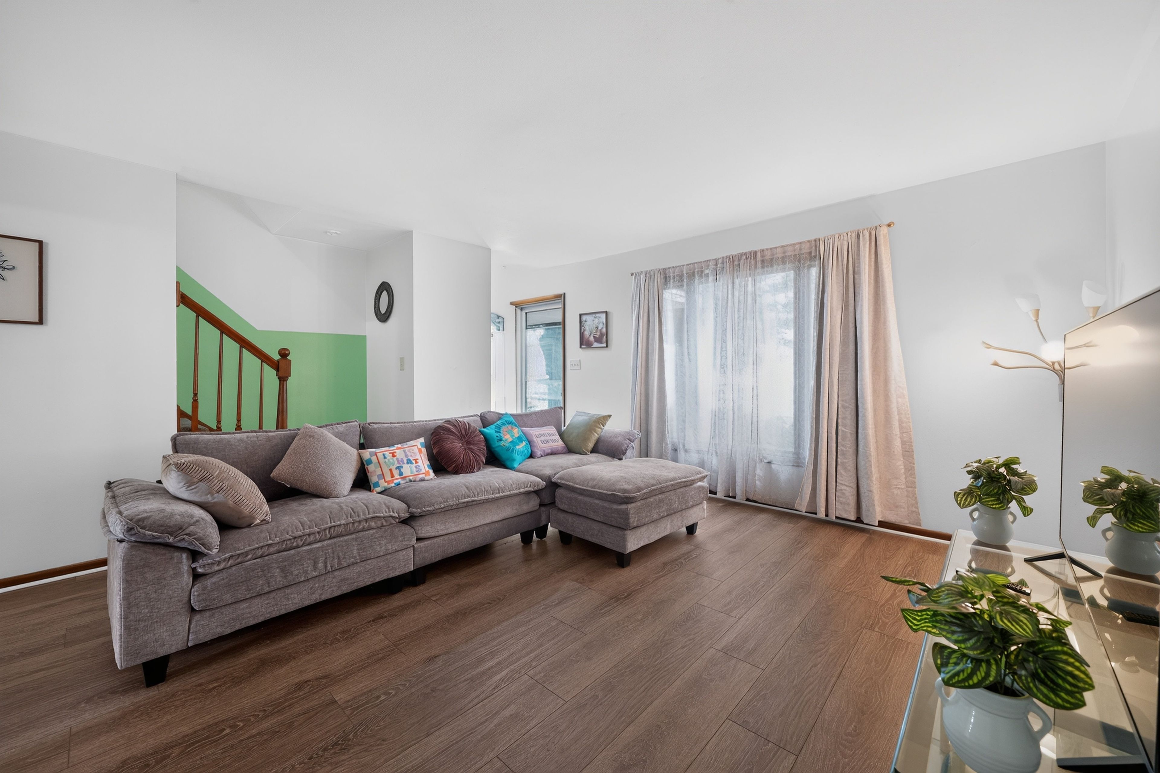 Un salon avec un canapé d’angle gris, un escalier agrémenté de touches de vert, une grande fenêtre avec des rideaux légers et un plancher en bois.