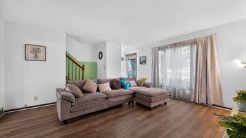 Un salon avec un canapé d’angle, un escalier et une grande fenêtre avec des rideaux.