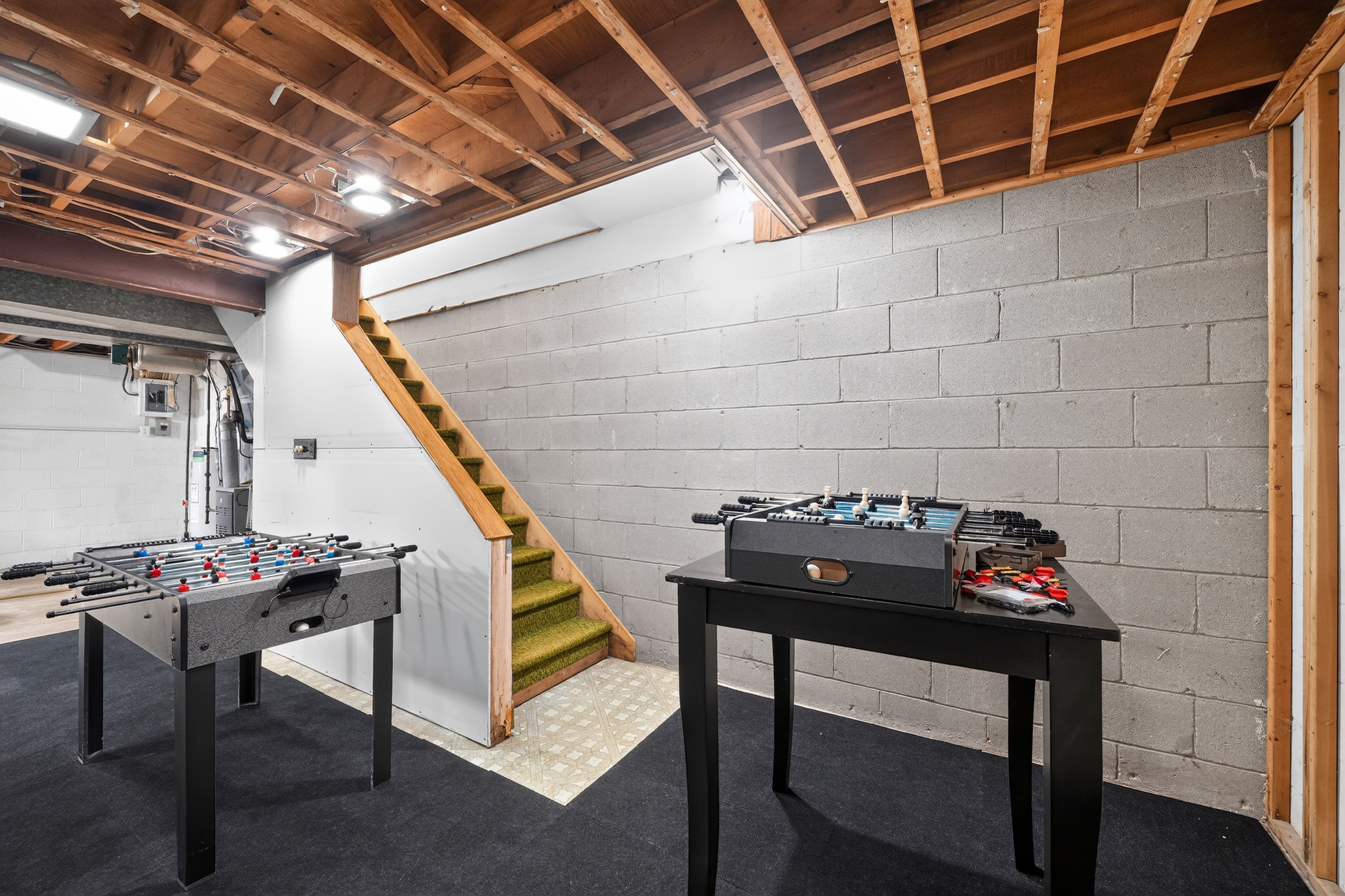 Une pièce avec deux tables de baby-foot, un escalier et des poutres en bois apparentes au plafond.