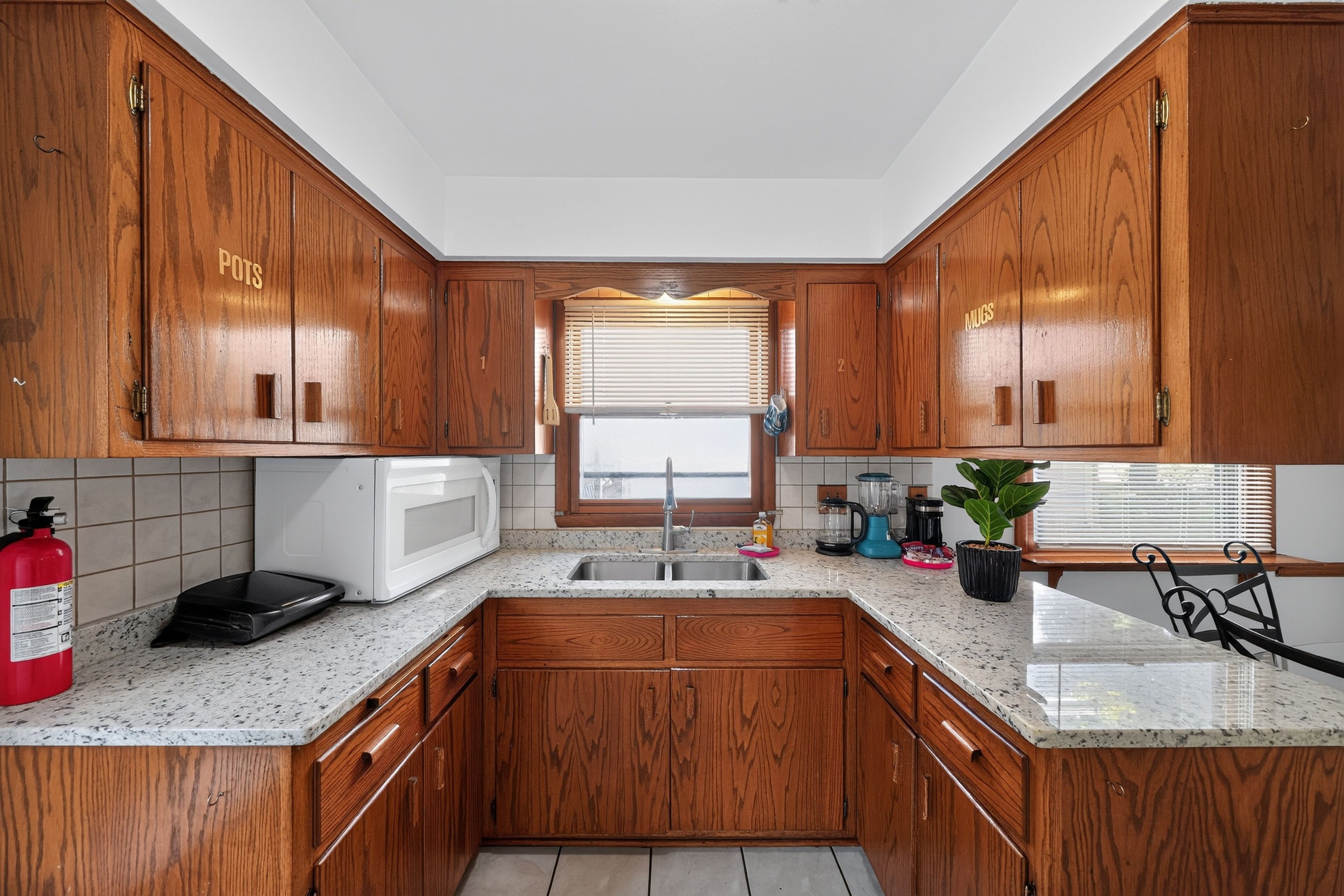 A kitchen with wooden cabinets, a microwave, a sink, and a window with blinds.