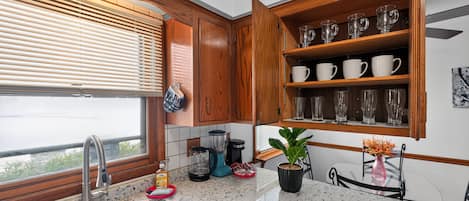 A kitchen with wooden cabinets, a granite countertop, a built-in shelf with glassware, and a window with blinds.