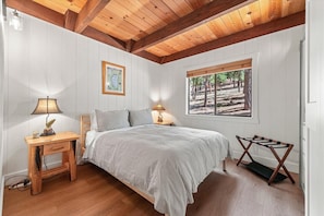 A bedroom with a bed, two bedside lamps, a small wooden nightstand, a window with a view of trees, and a wooden ceiling. - The Landing - Hanson 3 Bedroom Home (Truckee)